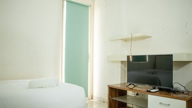 A hotel room with a bed, a TV on a wooden stand, a white desk, and a door with a teal frame. - Homey And Cozy Stay Studio At Margonda Residence 2 Apartment (Depok)