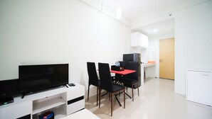 A modern, minimalistic room with a flat-screen TV, a dining table with four chairs, a small kitchenette, and a door leading to another room. - Best Choice And Cozy Living At 2Br At Bale Hinggil Apartment (Surabaya)