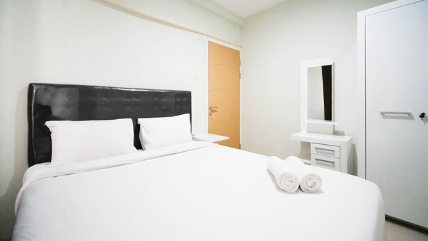 A neatly made bed with white linens and two rolled towels, a black upholstered headboard, a white bedside table with a mirror, and a white wardrobe. - Best Choice And Cozy Living At 2Br At Bale Hinggil Apartment (Surabaya)