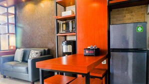A compact living space with a sofa, a wooden table with benches, a refrigerator, a microwave, and a coffee machine. - Modern And Best View 2Br Apartment At Bale Hinggil (Surabaya)