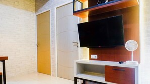 A modern living room with a flat-screen TV mounted on a wooden entertainment unit, a white-framed sofa, and a door with a circular handle. - Modern And Best View 2Br Apartment At Bale Hinggil (Surabaya)