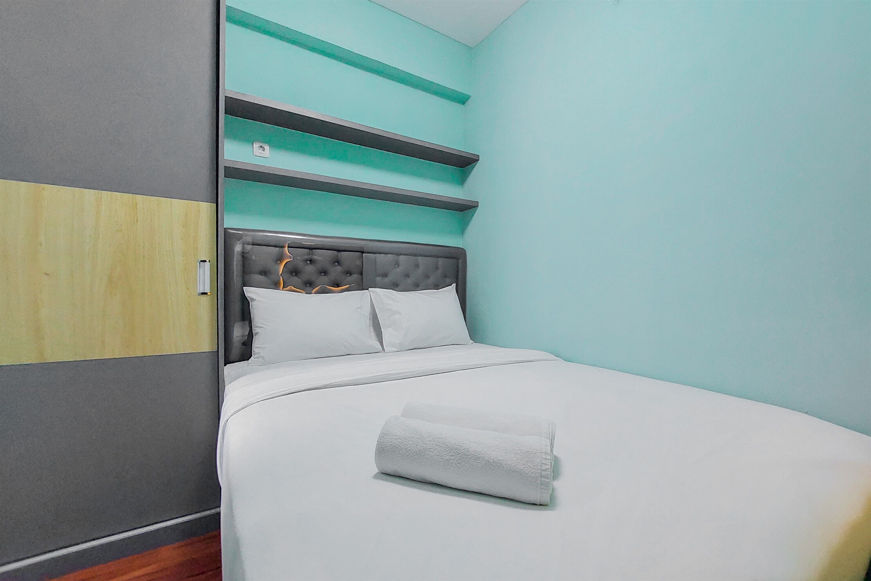 A neatly made bed with white linens and a rolled towel, a grey upholstered headboard, and teal-colored shelves above.