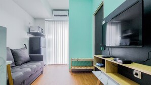 A modern living room with a grey sofa, a flat-screen TV, wooden shelves, and a window with blinds. - Nice And Simply Look 2Br At Serpong Garden Apartment (Cisauk)