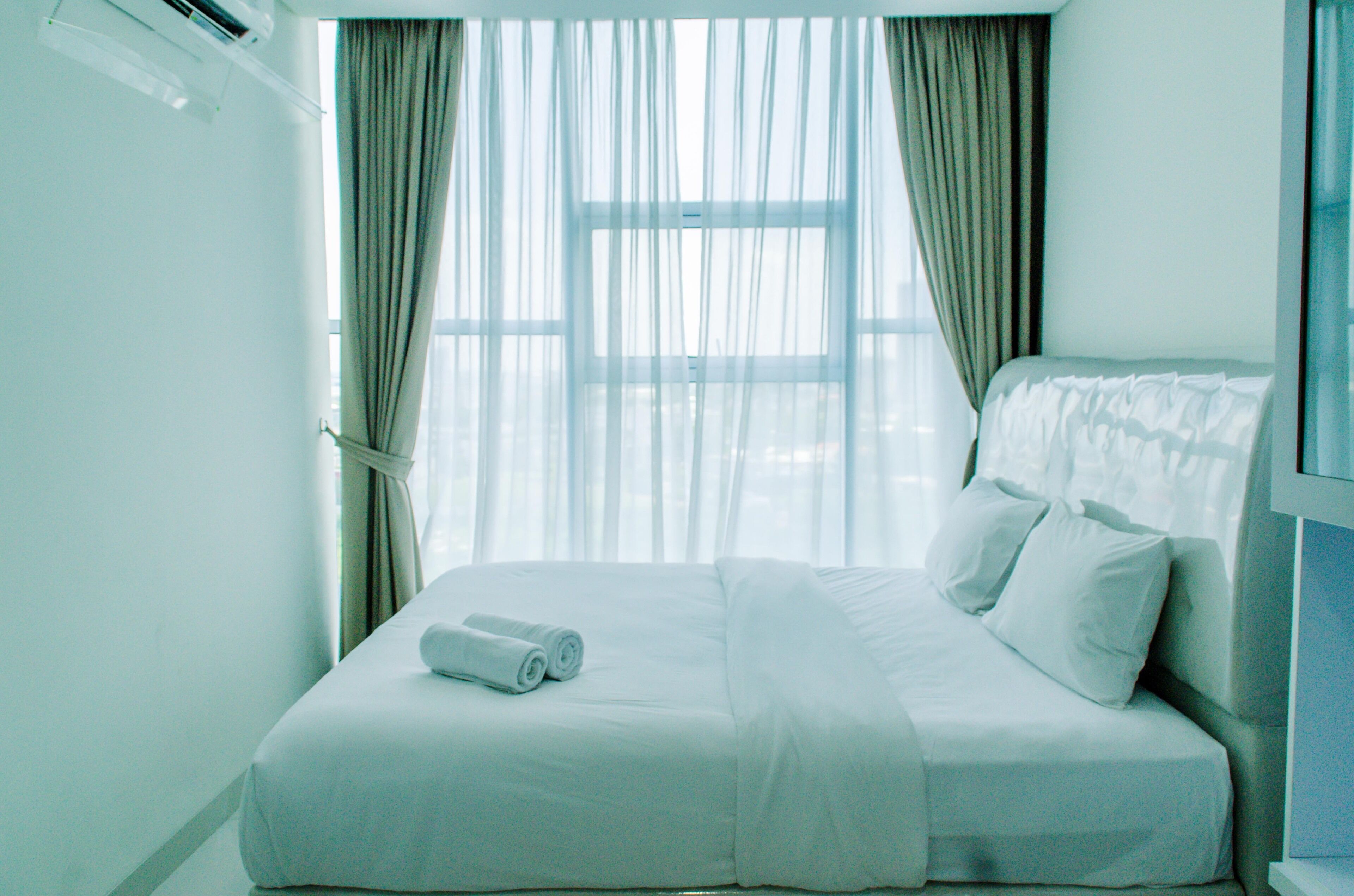 Una habitación de hotel con una cama grande, una ventana con cortinas translúcidas y una toalla enrollada sobre la cama.