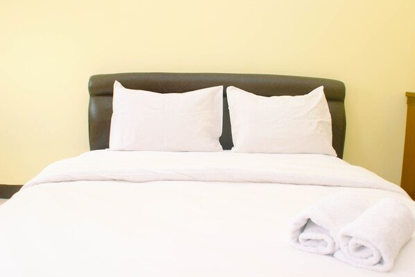 A neatly made bed with white linens and pillows, a rolled white towel on the side, and a dark headboard against a plain wall.