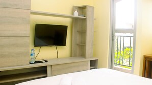 A hotel room with a bed, a TV mounted on a wooden shelf, a bottle of water, and a remote control. - Warm And Homey Studio Room At Maple Park Sunter Apartment (Jakarta)