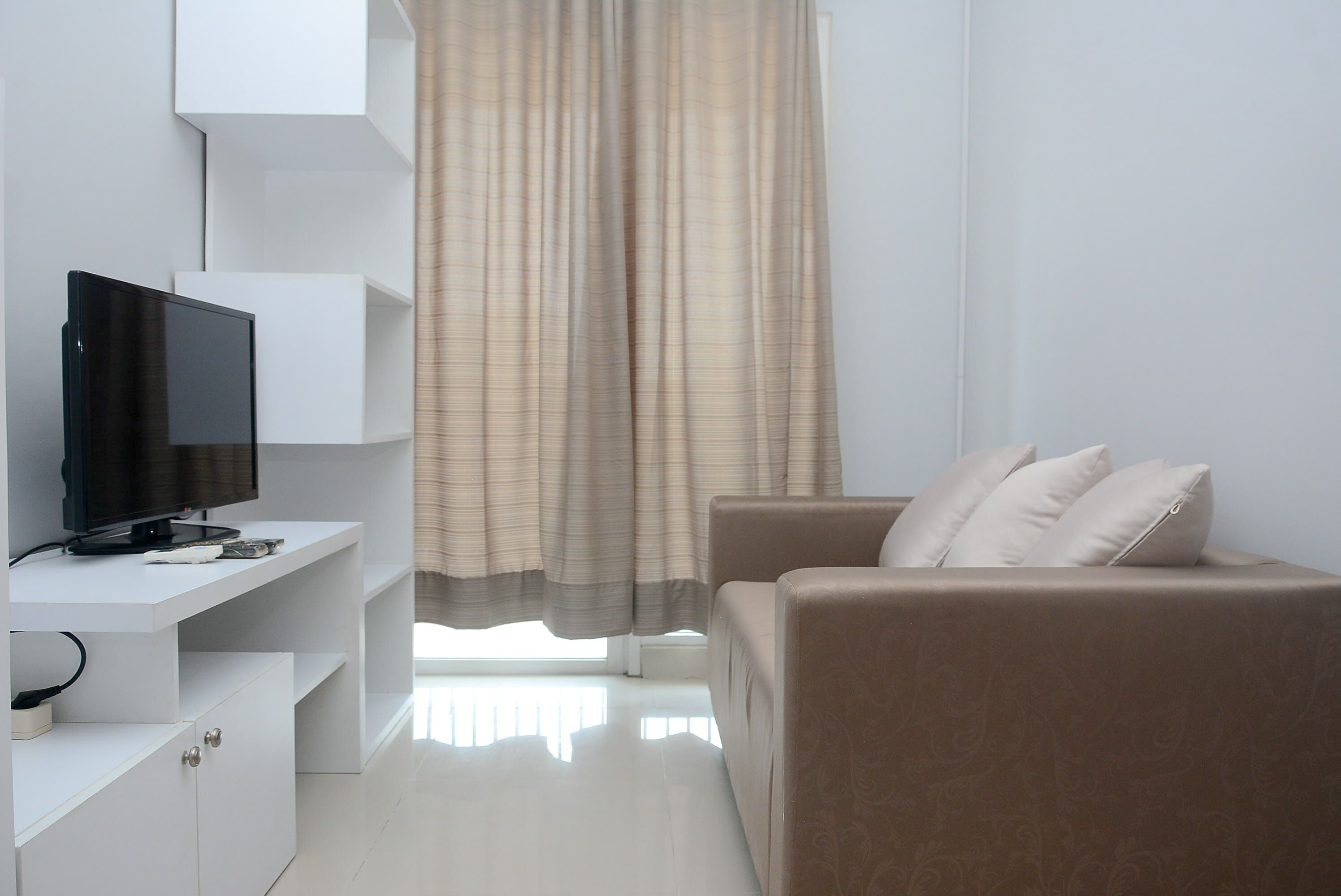 A modern living room with a flat-screen TV on a white console, a beige sofa with cushions, and a large window with curtains.