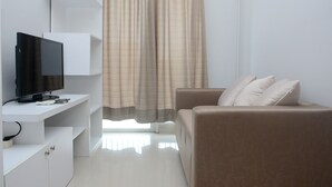 A modern living room with a flat-screen TV on a white console, a beige sofa with cushions, and a large window with curtains. - Unique And Fresh 1Br Signature Park Grande Apartment (Jakarta)