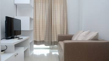 A modern living room with a flat-screen TV on a white console, a beige sofa with cushions, and a large window with curtains.