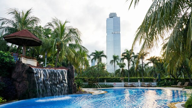A swimming pool with a waterfall feature, surrounded by palm trees and a view of a tall skyscraper in the background. - Elegant And Homey 2Br Apartment At Puri Casablanca (Jakarta)