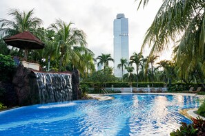 Piscina com cachoeira, cercada por palmeiras e vista para um arranha-céu ao fundo.