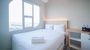 Habitació d'hotel amb una finestra gran, un llit amb roba de llit blanca, una taula de nit amb ampolla d'aigua i un coixí de viatge, i un taulell de nit de fusta.