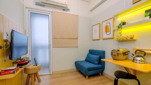A compact living space with a television, a sofa, a small table with a kettle, and a wall-mounted shelf with plants.