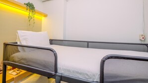 A single bed with a white mattress and a grey headboard, a small table with a bottle and a towel, and a hanging plant.