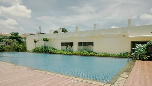 A swimming pool with a tiled deck, surrounded by greenery and a building with large windows.