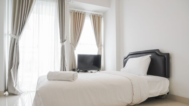 A neatly made bed with a flat-screen TV and a pillow, a window with curtains, and a clean, tiled floor.