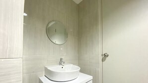 A modern bathroom with a white sink, a round mirror, and a door with a handle. - Homey And Elegant Studio Vasanta Innopark Apartment (Cikarang)