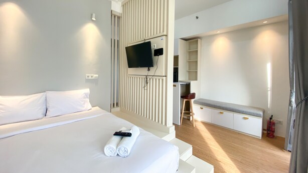 A modern hotel room with a large bed, a TV mounted on a wall, a small kitchenette, and a seating area with a bench and chair. - Homey And Elegant Studio Vasanta Innopark Apartment (Cikarang)
