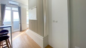 A room with a wooden floor, a white door, a white wall with a vertical panel, and a window with curtains. - Homey And Elegant Studio Vasanta Innopark Apartment (Cikarang)