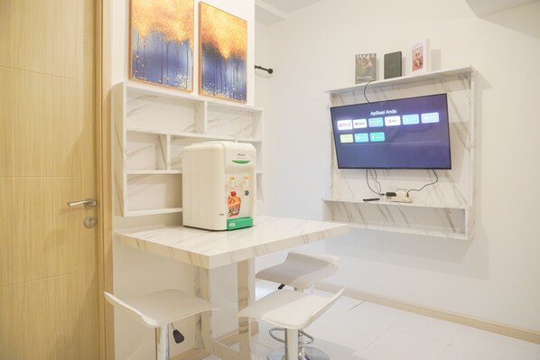 A modern room with a white desk, a coffee machine, a TV mounted on the wall, and a shelf with books and decor.