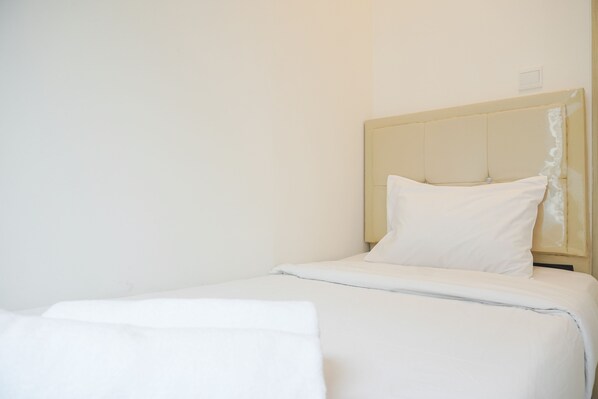 A neatly made bed with white linens in a simple room with a plain wall and a wooden headboard.