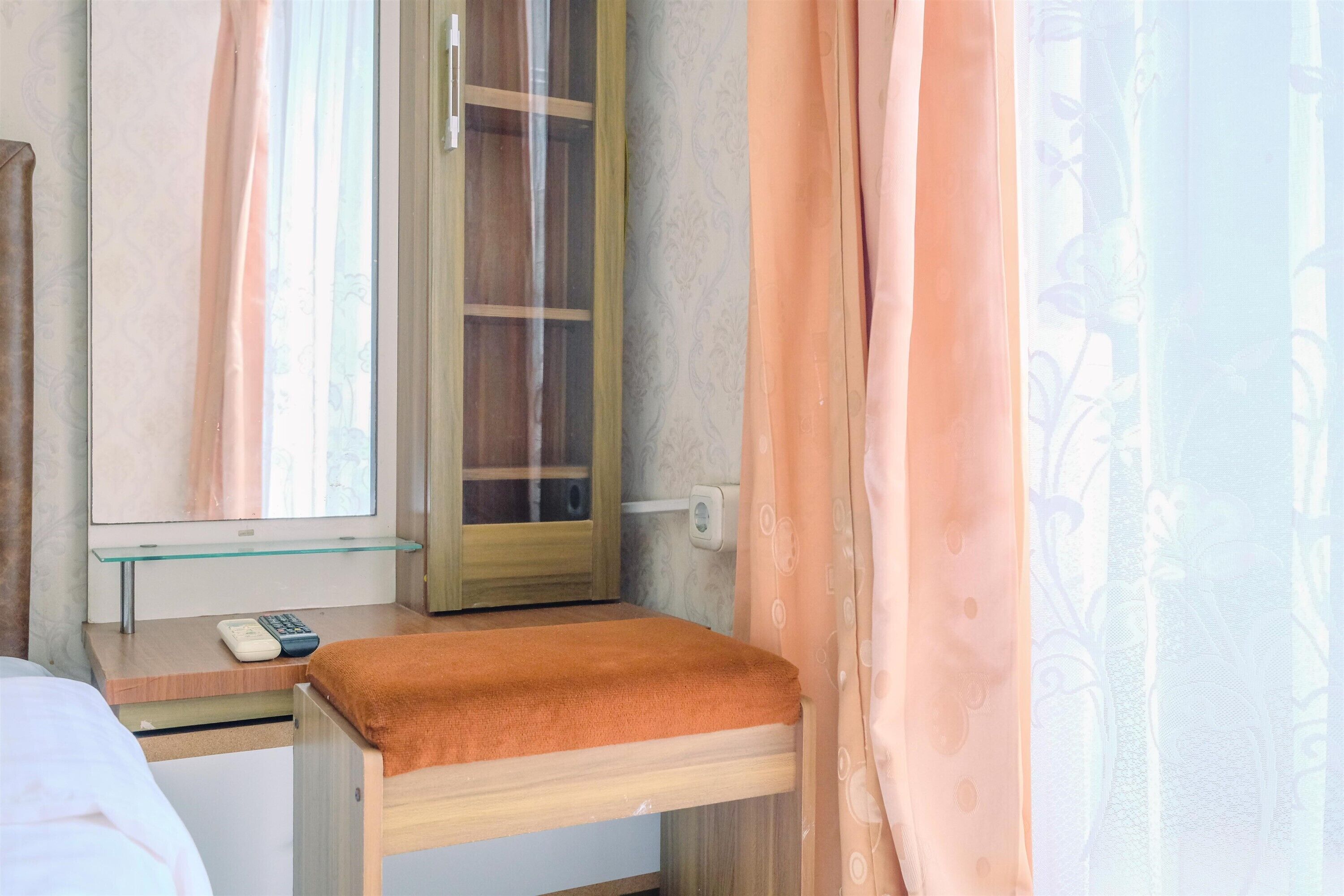 A hotel room with a bed, a bedside table with a remote control, a glass cabinet, and a window with curtains.
