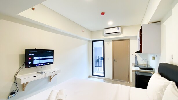 A hotel room with a bed, a TV mounted on a wall, a desk with a computer, and a door leading to a balcony. - Homey And Simply Look Studio Meikarta Apartment (Cikarang)