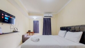 A hotel room with a bed, a TV mounted on the wall, an air conditioning unit, and a small table with a chair. - Cozy And Minimalist Studio Great Western Apartment (Tangerang)