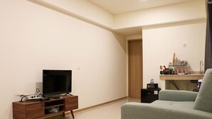 Una sala de estar con una televisión de pantalla plana sobre un mueble de madera, un sofá gris y una zona de cocina con un escurreplatos y armarios.