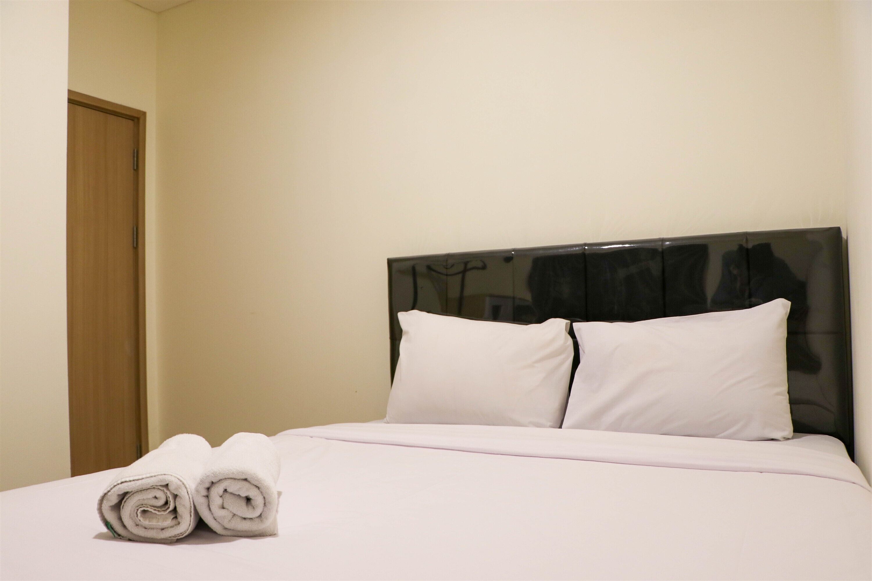 Una cama bien hecha con sábanas y almohadas blancas, una puerta de madera a la izquierda y una pared lisa.
