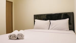 Una cama bien hecha con sábanas y almohadas blancas, una puerta de madera a la izquierda y una pared lisa.