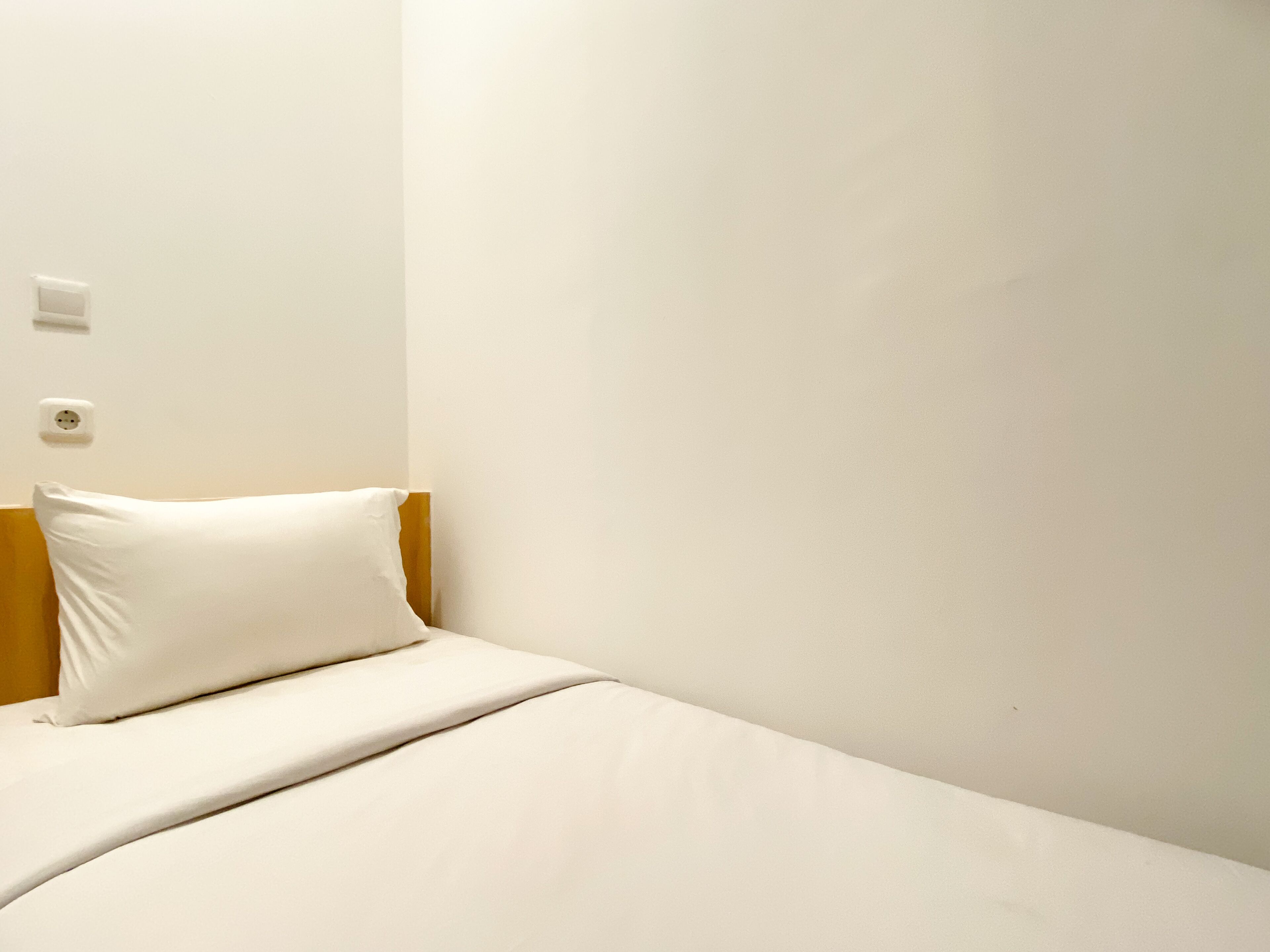 A single bed with a white pillow and a wooden headboard in a room with plain white walls and a visible electrical outlet.