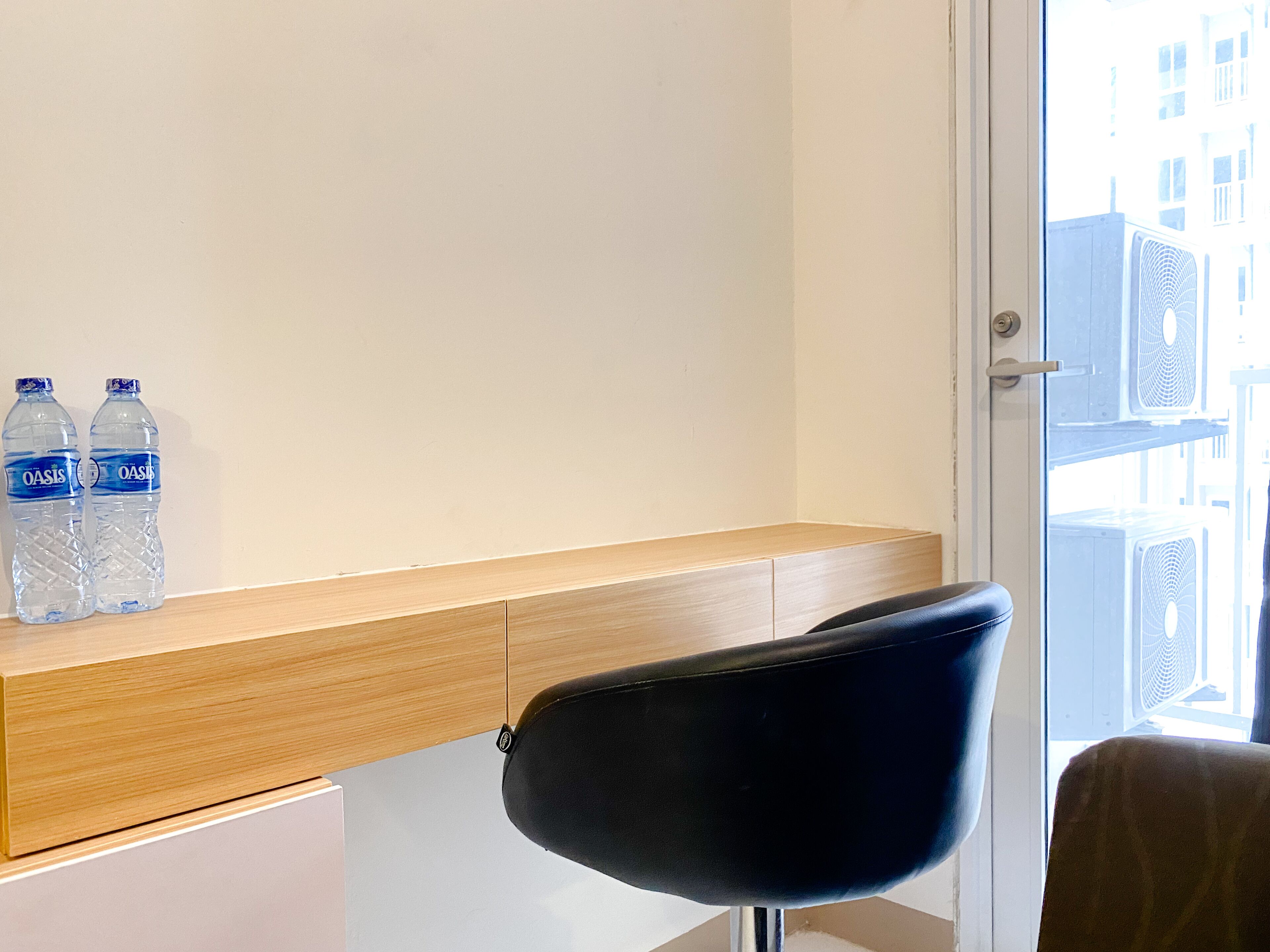 A hotel room with a wooden desk, two water bottles, a black chair, and a window with air conditioning units outside.