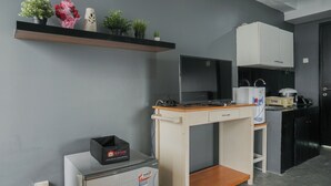 A compact kitchenette with a built-in oven, a small refrigerator, and a microwave. There is a shelf above the counter holding potted plants and a vase with flowers. - Nice And Warm Studio At Belmont Residence Puri Apartment (Jakarta)