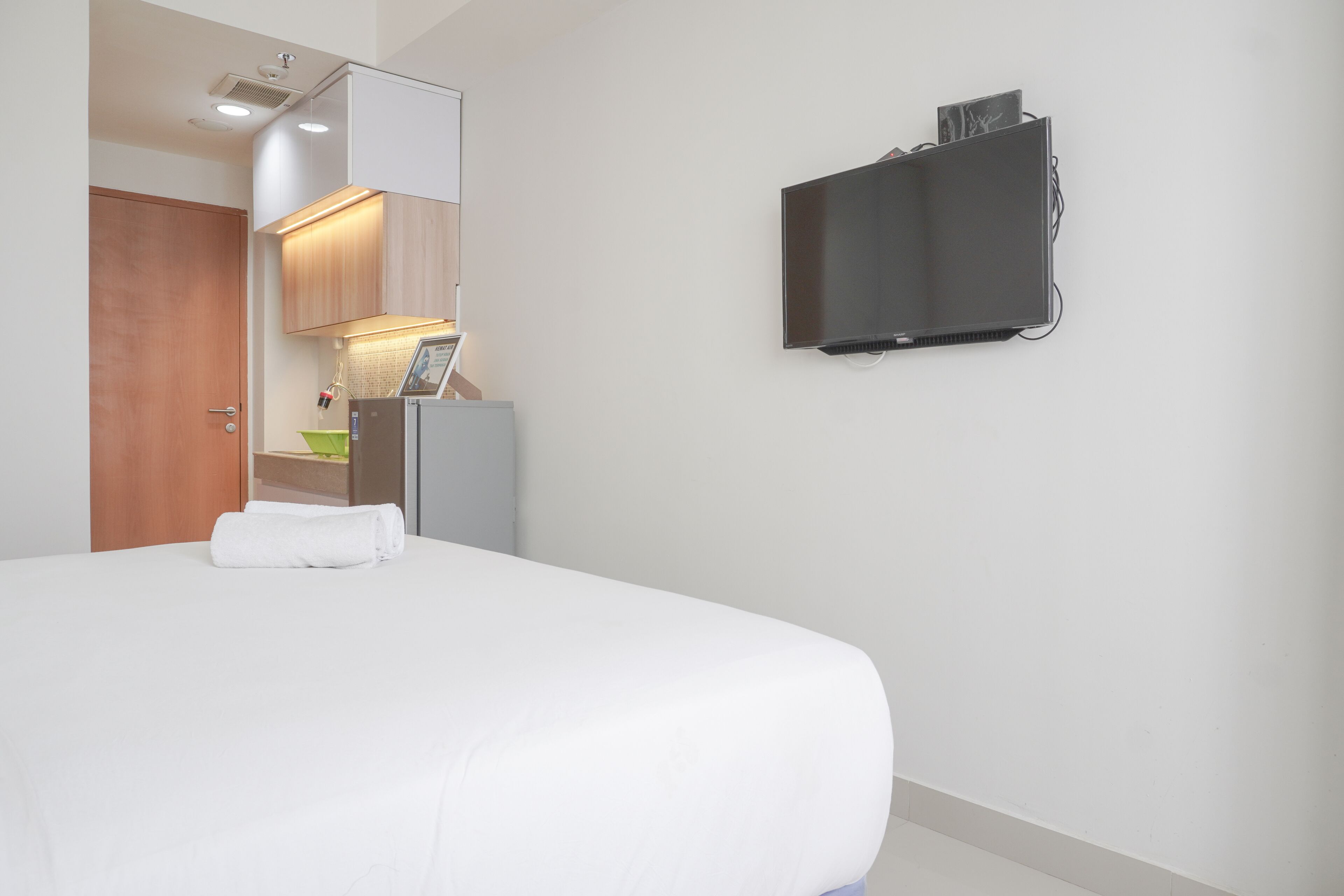 A hotel room with a bed, a flat-screen TV mounted on the wall, a wooden door, and a kitchenette area with a refrigerator and a small table.