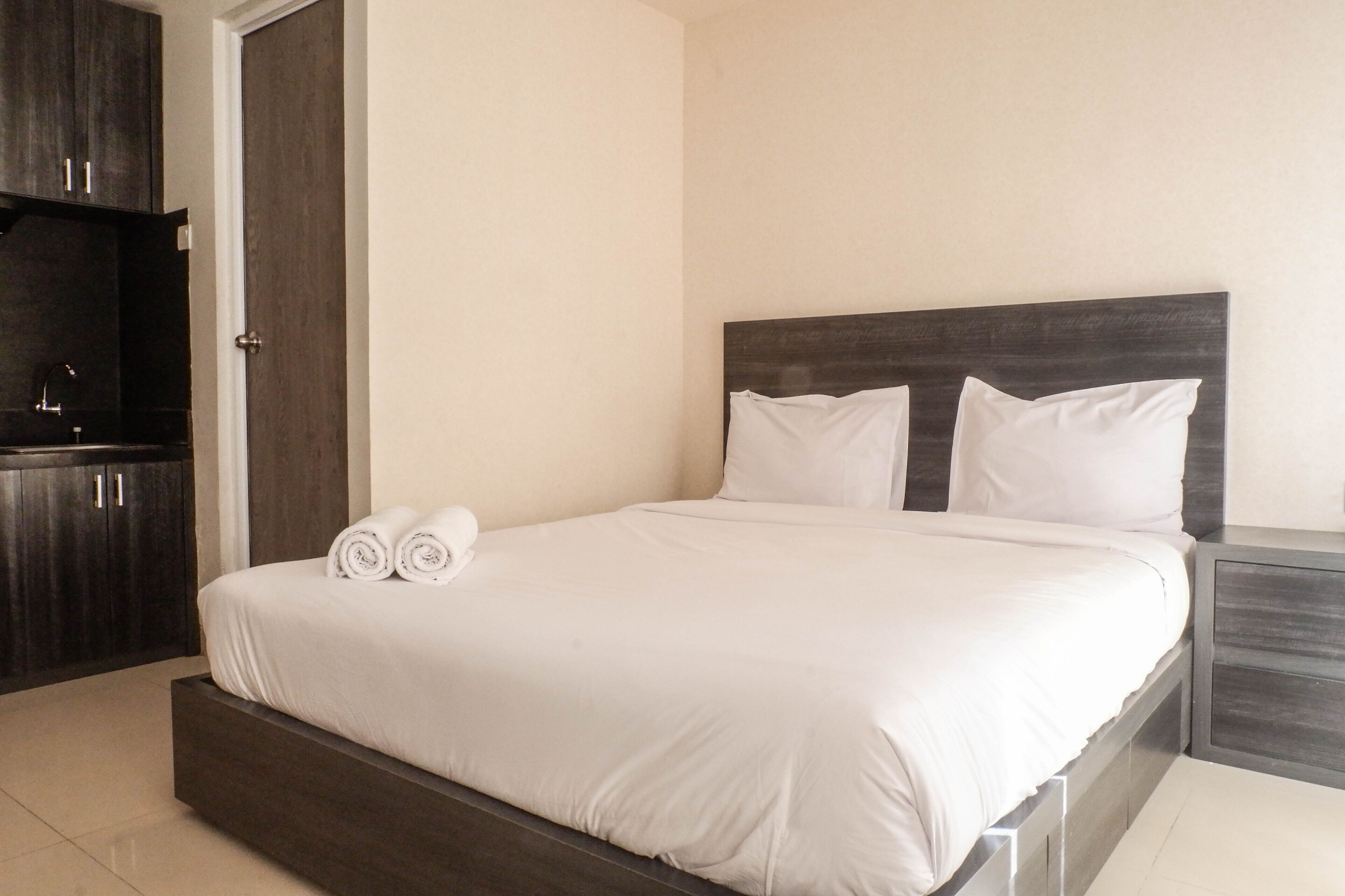 Una cama bien tendida con sábanas blancas y dos almohadas, un cabecero de madera oscura y una mesita de noche con un cajón.