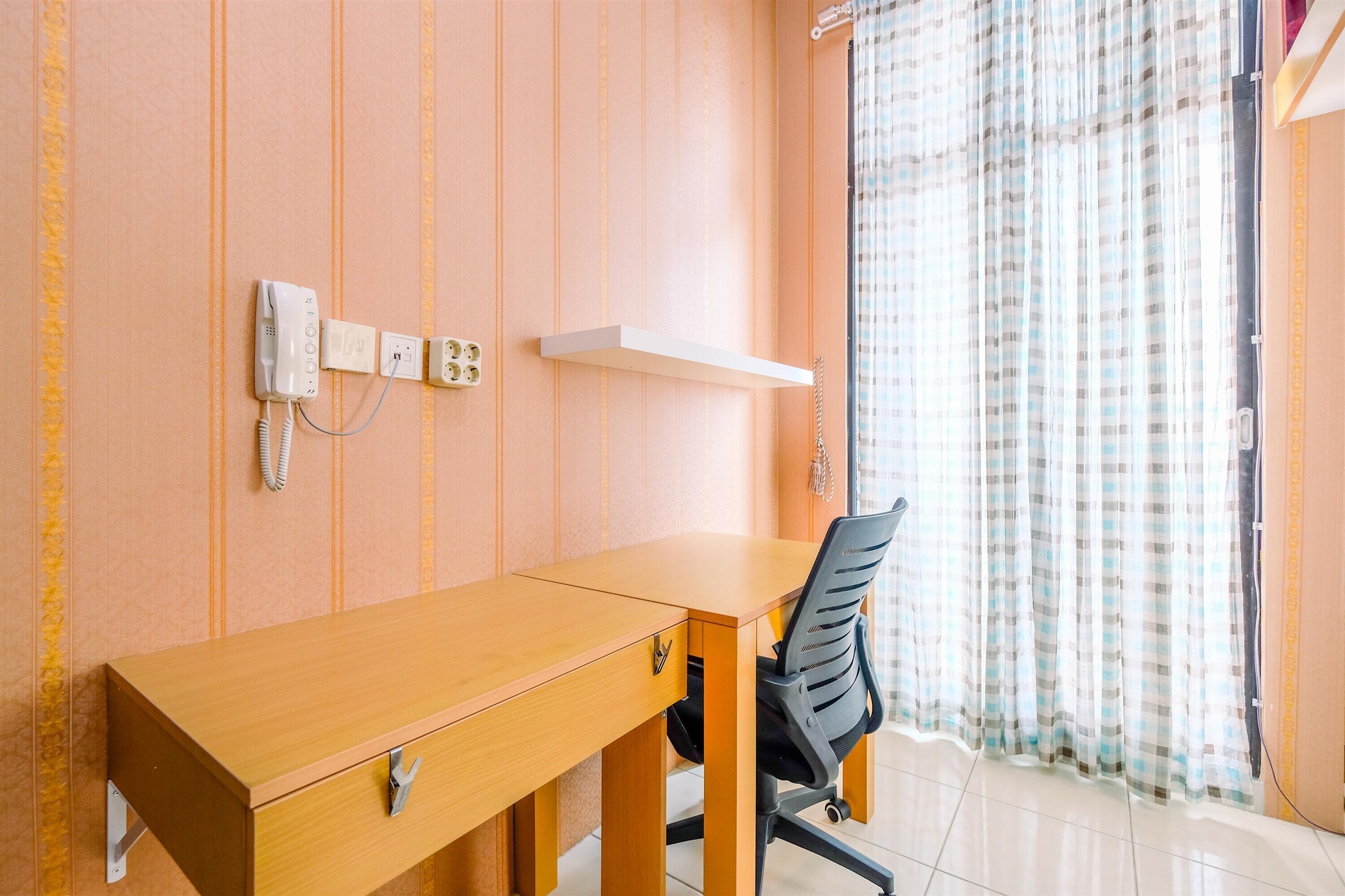 Ein Zimmer mit einem Holztisch, einem Stuhl, einem Telefon und einem an der Wand befestigten Regal.