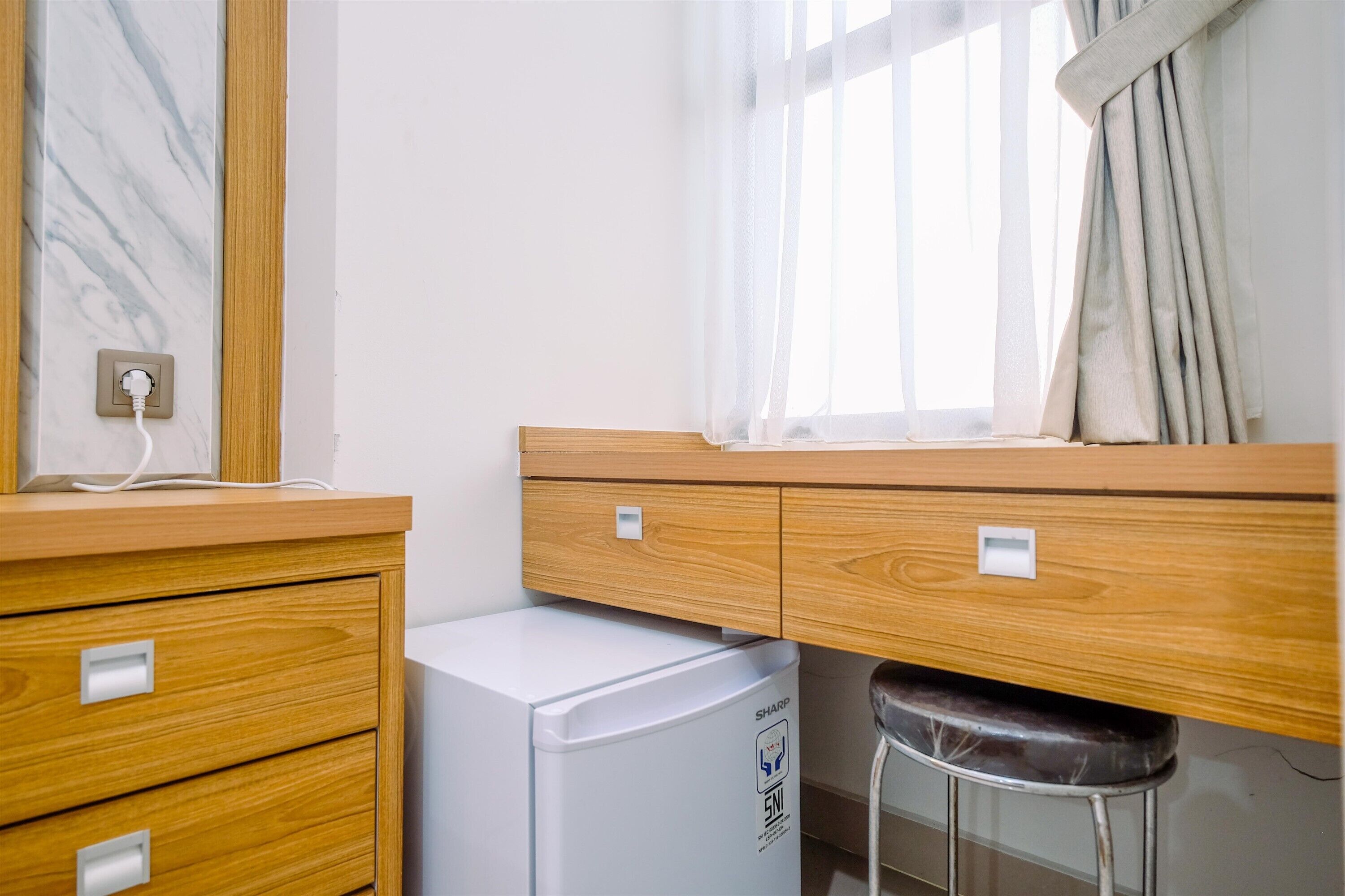 A compact room with a wooden desk, a small white refrigerator, a stool, and a window with sheer curtains.