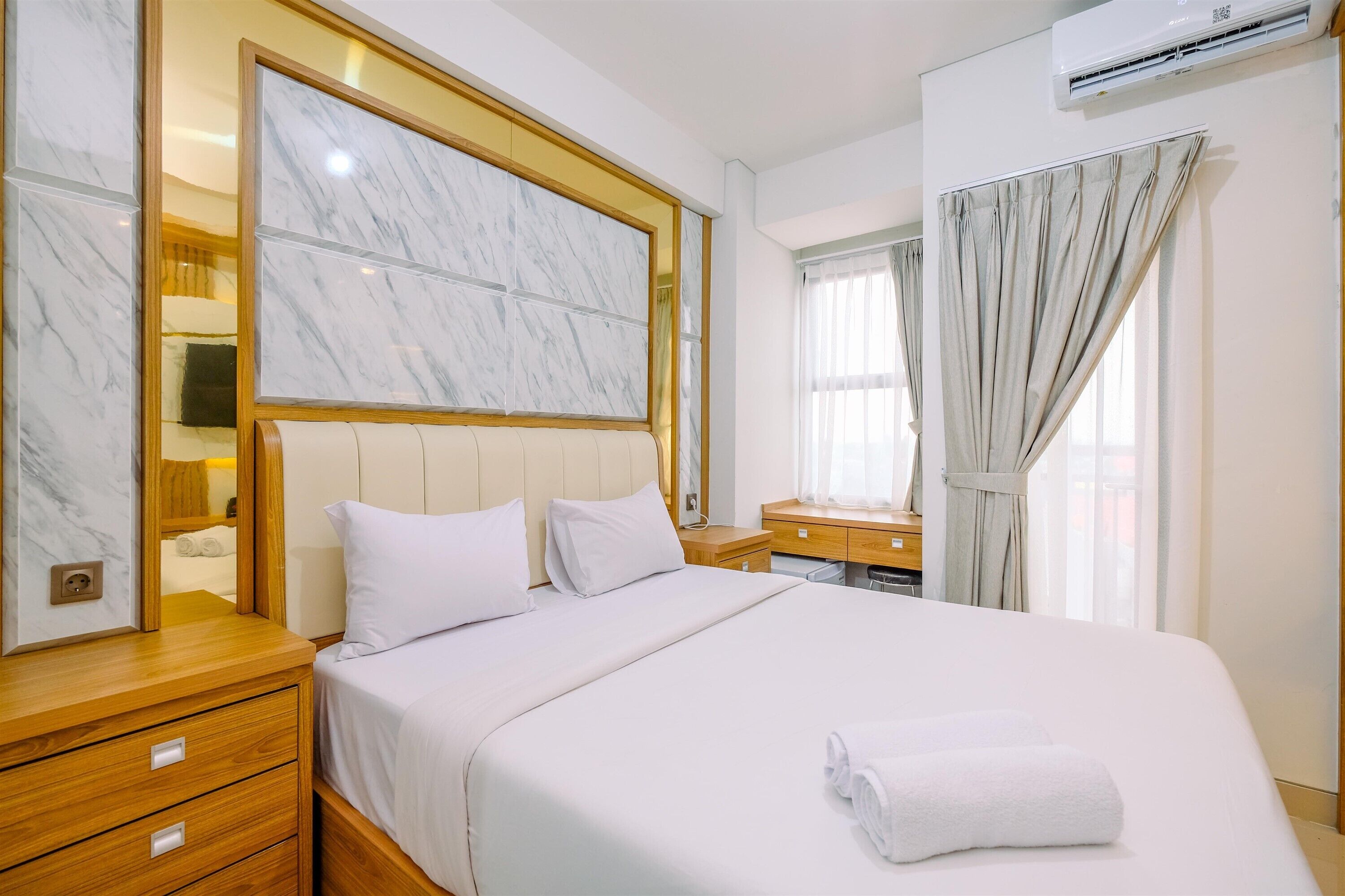A hotel room with a large bed, wooden nightstands, a marble headboard, a mirror, and a window with curtains.