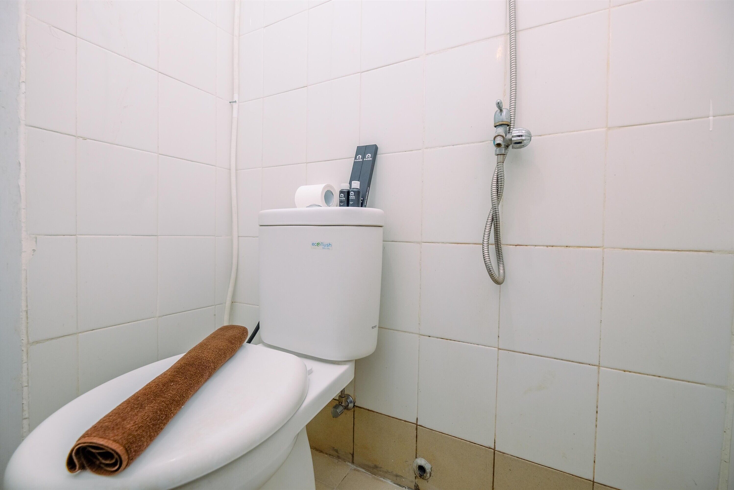 A bathroom with a white toilet, a brown towel on the seat, a toilet paper holder, and a shower hose.