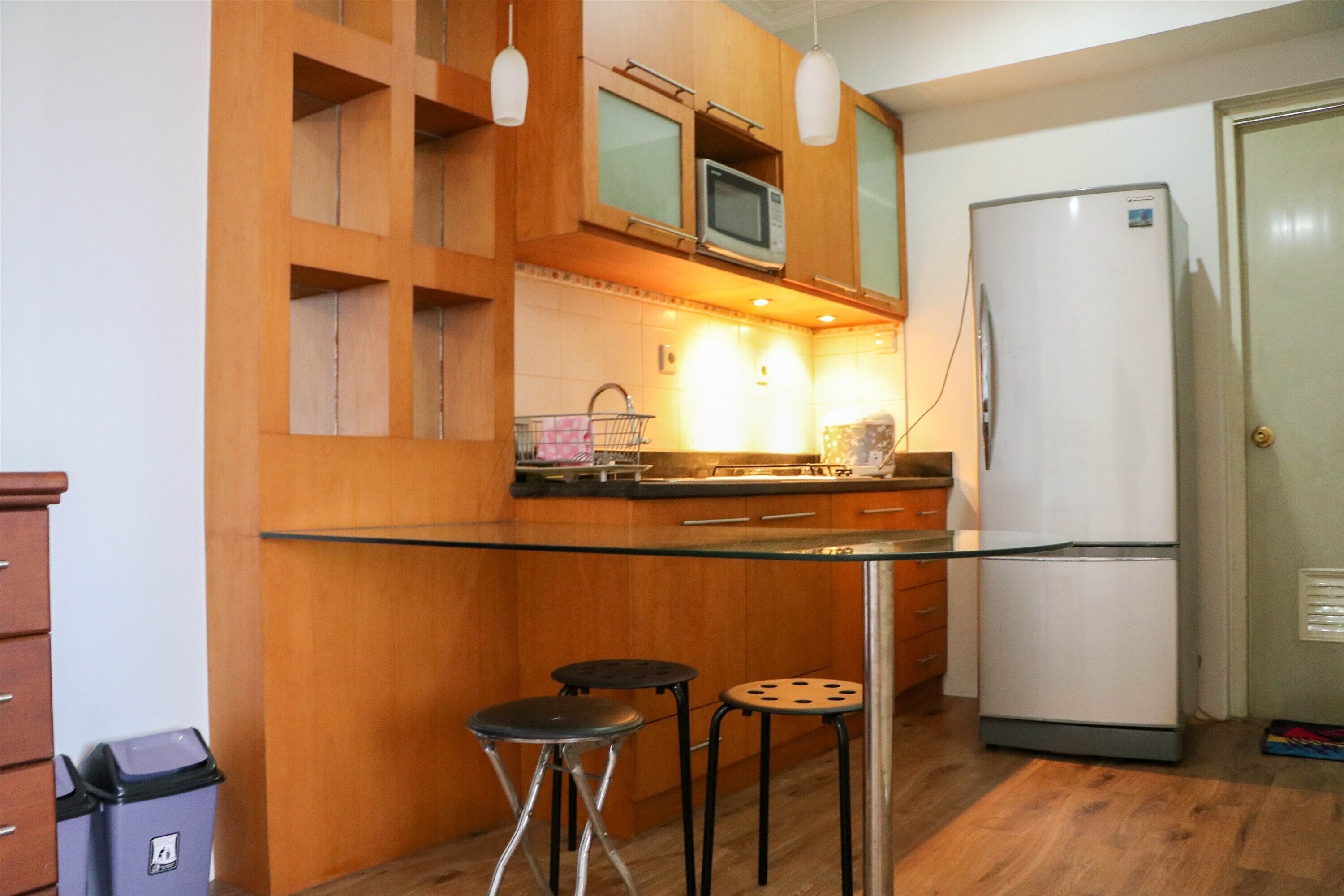 A compact kitchen with wooden cabinets, a microwave, a refrigerator, and two stools.