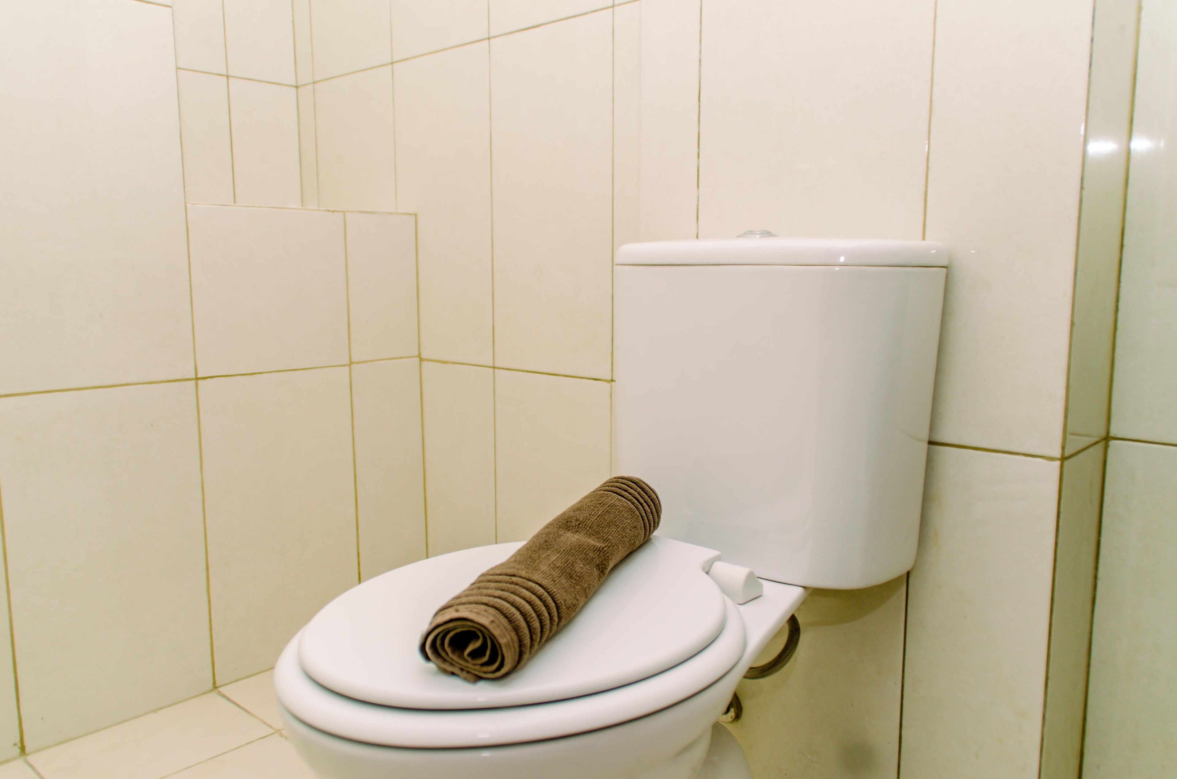 A toilet with a rolled-up towel on the lid in a bathroom with white tiled walls.