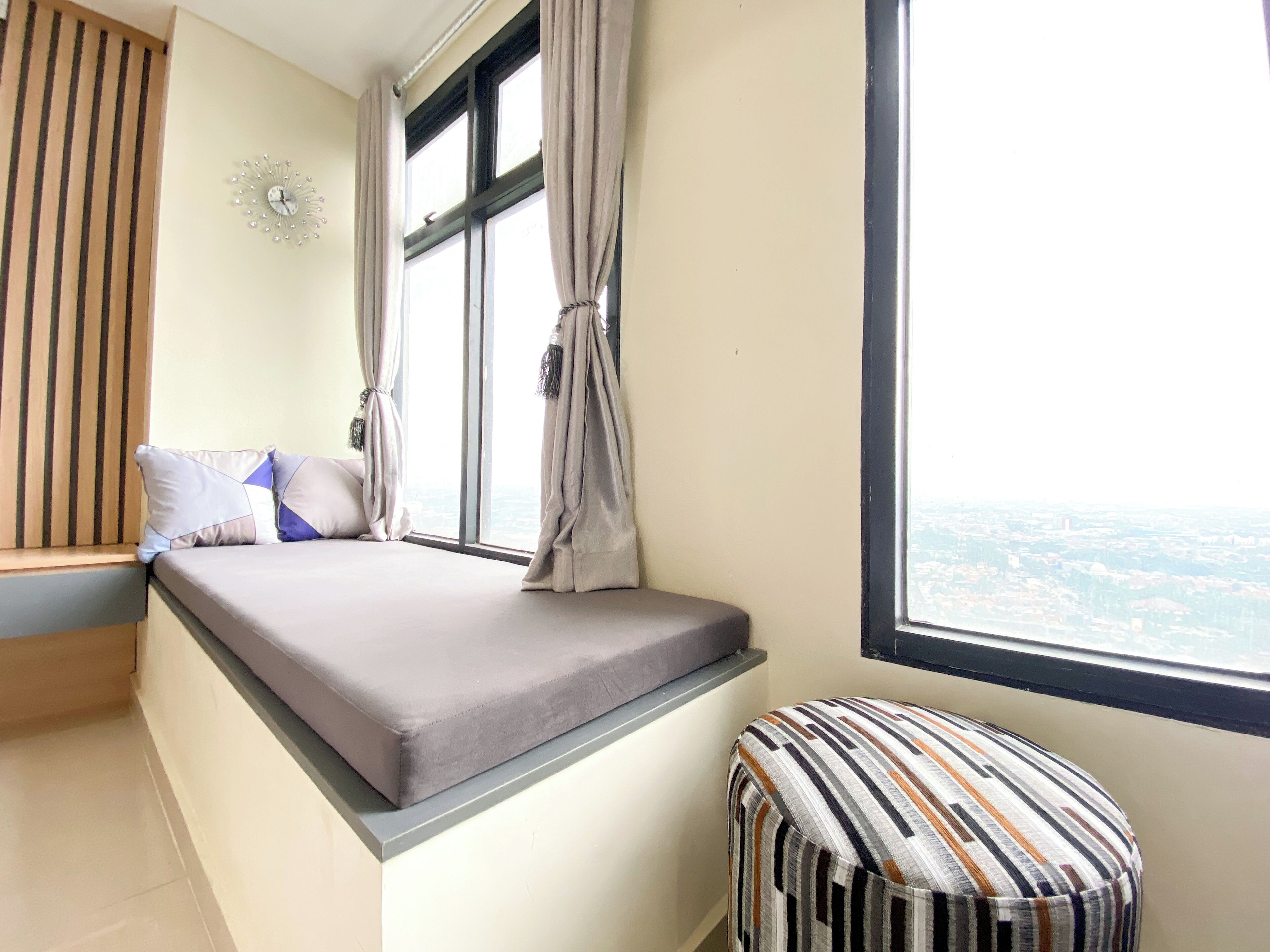 Un dormitorio con una cama, una ventana con cortinas, un reposapiés a rayas y un reloj de pared.