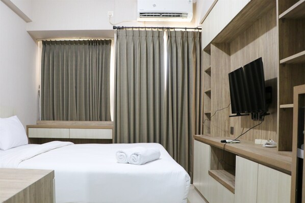 A hotel room with a bed, a TV mounted on a wooden panel, and a window with curtains. - Elegant And Nice Studio Room At Citra Living Apartment (Jakarta)
