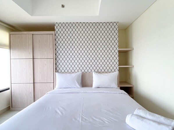 A bedroom with a bed, two pillows, a headboard with a geometric pattern, a wardrobe, and a shelf unit. - Simply Look Studio At Pollux Chadstone Apartment (Cikarang)