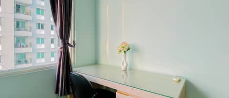 Una habitación con un escritorio de vidrio, una silla negra, un jarrón con flores y vista a un edificio con balcones.