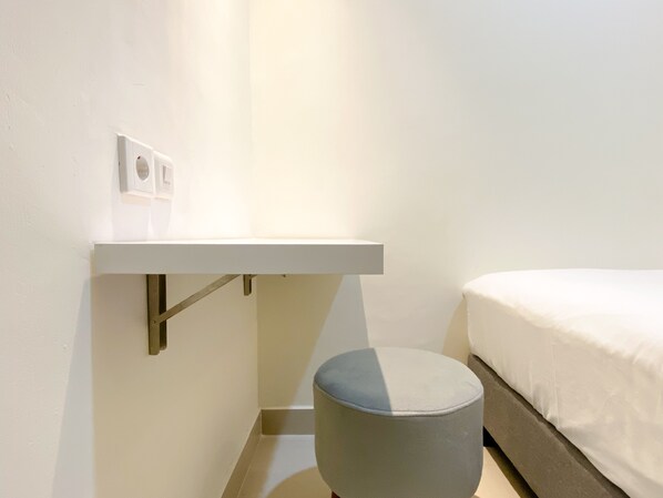 A minimalist room with a bed, a small round ottoman, a white shelf, and a wall-mounted light fixture. - Comfort Stay And Homey 2Br Pollux Chadstone Apartment (Cikarang)