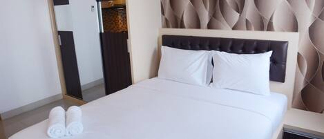 A neatly made bed with white linens and two rolled towels, a tufted headboard, and a mirror on the wall.