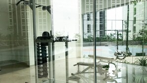 A gym with exercise equipment, large windows, and a view of a building and greenery outside. - Nice And Cozy Studio At Pollux Chadstone Apartment (Cikarang)