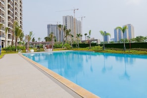 A swimming pool with a paved walkway, surrounded by greenery and palm trees, with high-rise buildings and construction cranes in the background.